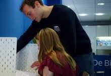 Man assisting a woman with forms at a desk in a service area, leaning over to review documents together on a light desk panel beside them.